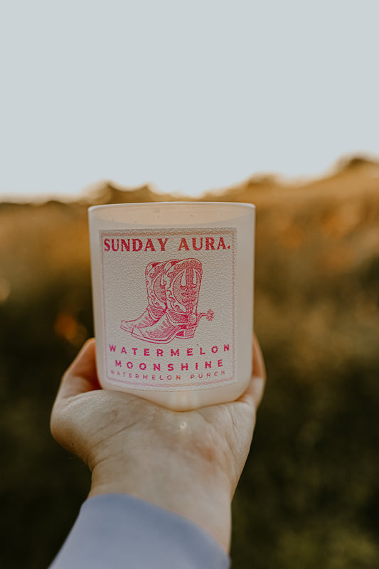 Hand holding a 'Sunday Aura' candle with a boot design against a blurred natural background
