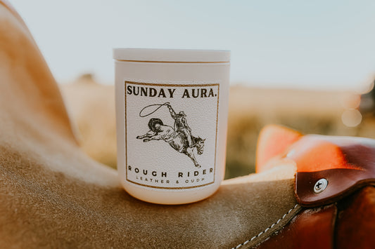Candle labeled 'Sunday Aura Rough Rider' on a leather surface with a blurred natural background