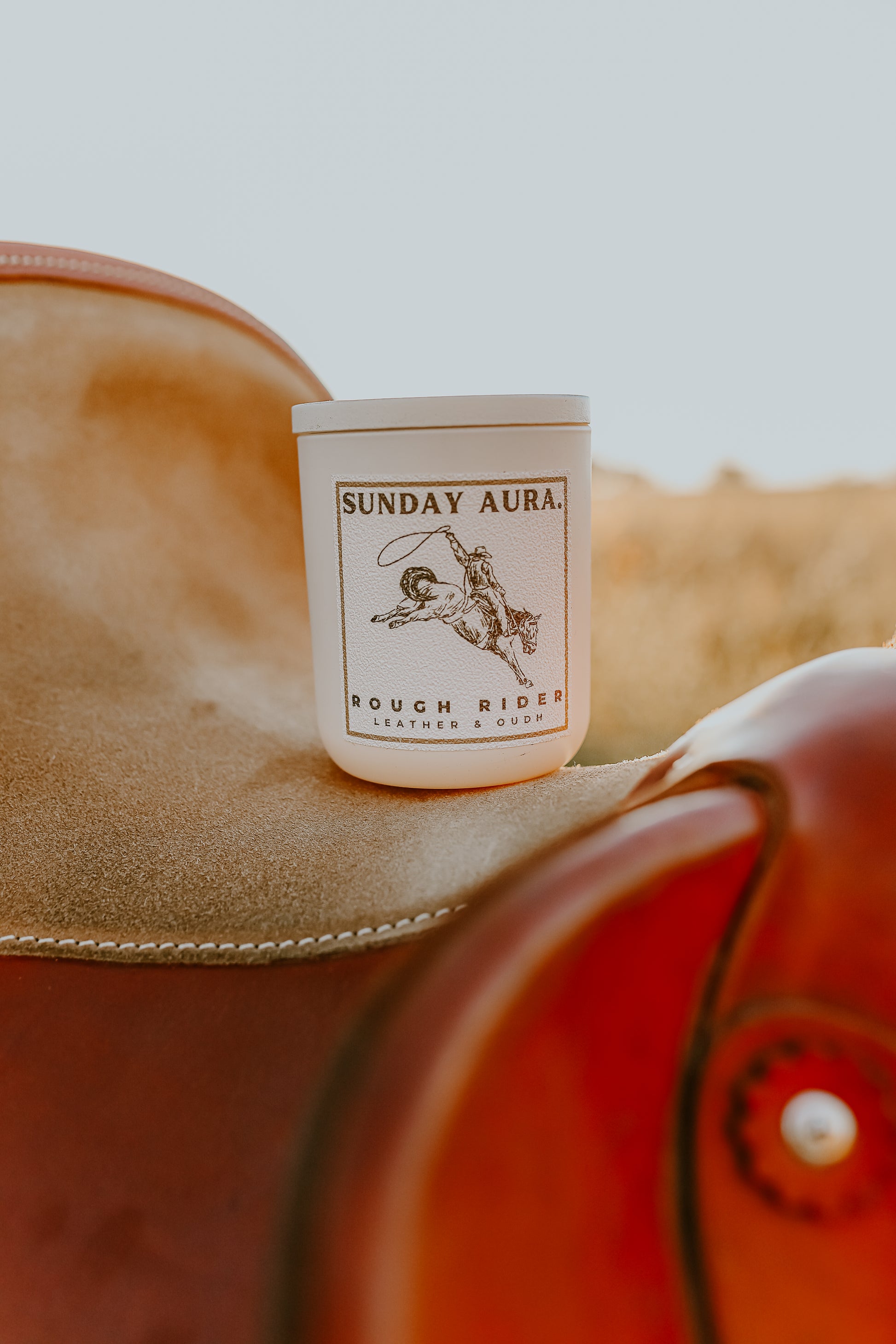 Candle labeled 'Sunday Aura Rough Riders' on a leather surface with a blurred background