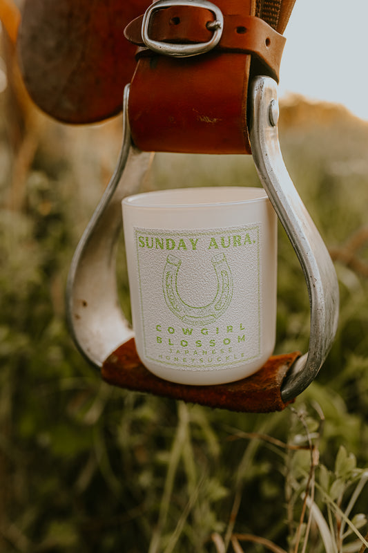 A candle with a label that reads 'SUNDAY AURA COWGIRL BLOSSOM', held in a metal cup, with a background of a horse and natural elements.
